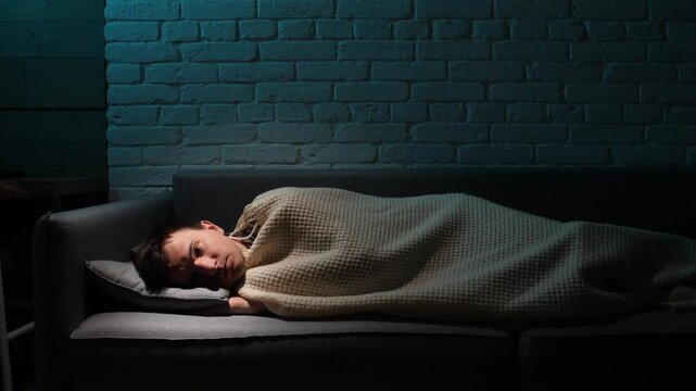 Man lying awake on a couch late at night under a blanket, restless and pensive, struggling with insomnia, anxiety and sadness in a dim, lonely room