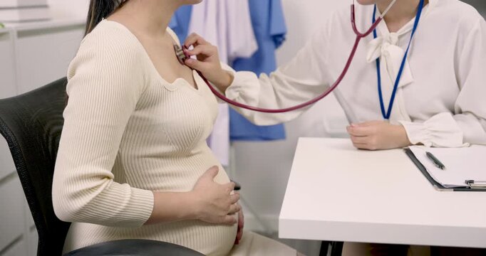 Obstetrician examining heartbeat of pregnant Asian woman with stethoscope during prenatal consultation in clinic showing medical attention trust and care during third trimester maternity checkup