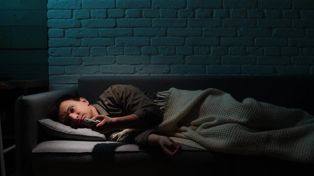 Man lying awake on couch in a dark living room late at night, clutching a remote, restless and lonely, battling insomnia, boredom and quiet despair under the tv glow