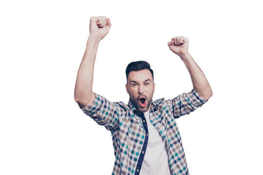 Amazed brunet guy with bristle in casual checkered shirt celebrating victory of favorite sport team with raised arms, cams,  shouting, screaming, yelling with open mouth on grey background