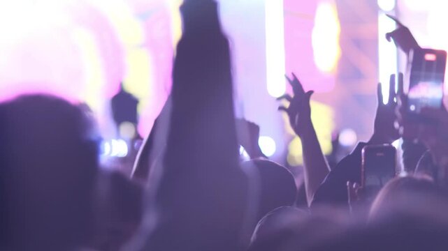 Large and dense crowd of people dancing on dance floor in front of stage. People raise hands up and rhythmically energetically dance jumping and having fun to electronic music and light effects.