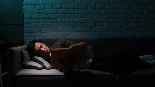 Young man dozing on a gray sofa in a dim living room, falling asleep with an old hardback book in hand, soft lamp light creating a calm, cozy evening atmosphere
