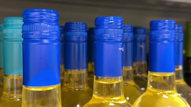 Rows white wine bottles blue caps closeup on shelf, golden liquid in clear glass, assorted brands with visible labels, screw top