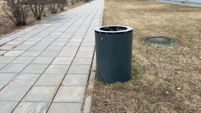 Grey waste bin on paved path, small metal receptacle stands beside dry spring grass and tiled sidewalk, scattered leaf litter
