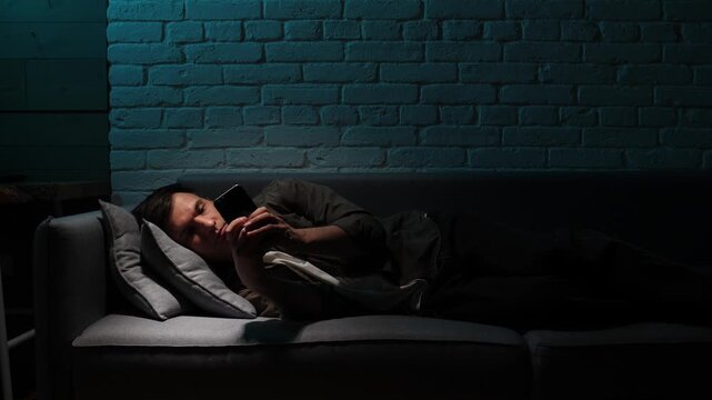 Man lying on a sofa at night, scrolling through content on a smartphone, with the blue screen light illuminating his face, causing sleep problems and digital addiction