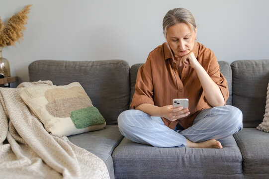 Thoughtful middle aged woman using smartphone while sitting on sofa, making a decision and comparing options online. Modern lifestyle and choice concept.