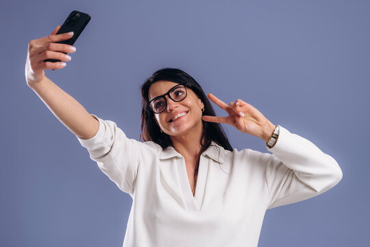 Woman with long dark hair wearing glasses takes a selfie with a smartphone, smiling and making a peace sign against a solid purple background