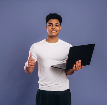 Young male student with curly hair holds a laptop in one hand and gives a thumbs up with the other against a solid purple background