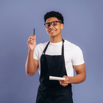 Young African American male barista wearing black apron holds a pen and notepad while smiling against a blue background in a coffee shop setting