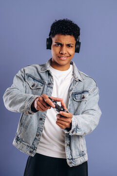 Young male gamer wearing headphones holds a video game controller while posing against a solid blue background, showcasing a casual style and focused expression