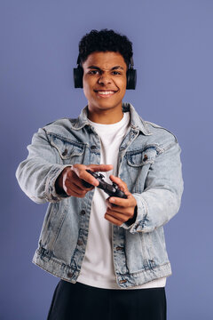 Young male gamer wearing headphones holds a video game controller while smiling against a solid purple background, showcasing a casual gaming lifestyle