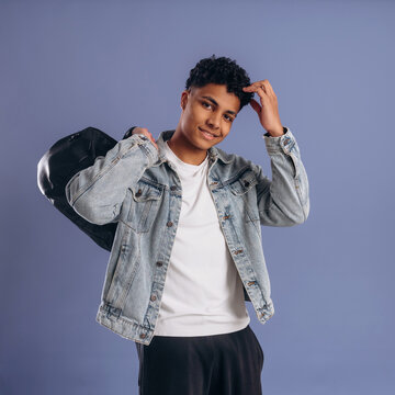 Young male wearing denim jacket and white t-shirt poses confidently with a black bag over his shoulder against a solid purple background