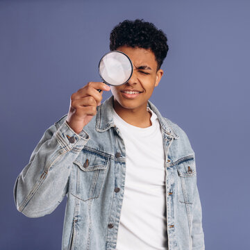 Young African American male wearing denim jacket holds magnifying glass to his eye, posing against a solid purple background in a playful and inquisitive manner