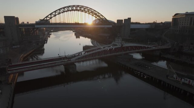 Cinematic sunrise over Newcastle showing the sun rising behind the Tyne Bridge, with the Swing Bridge and Millennium Bridge reflected in the river.