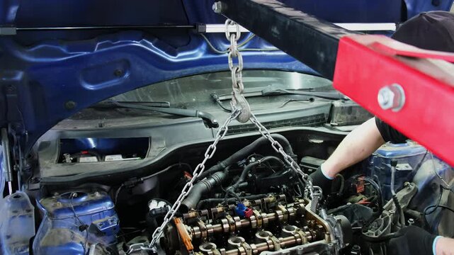 Worker dismantles car engine with hanging on hoist chain in repair shop slow motion. Mechanic lifts motor with hydraulic machine to fix at station