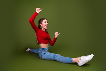 Naklejka premium Young stylish woman in red crop top and jeans performs a joyful split against a green studio backdrop