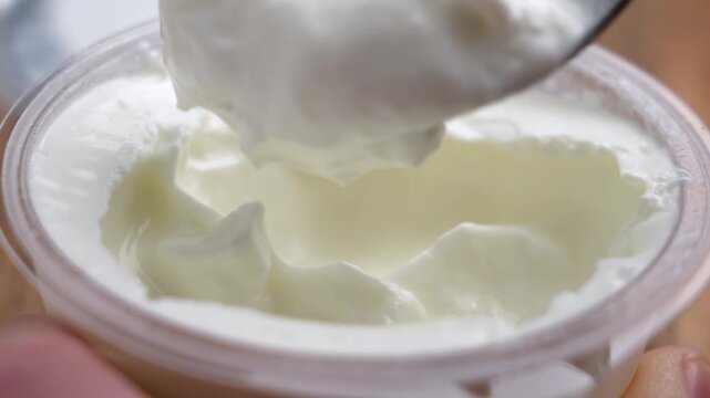 Metal spoon scooping thick yogurt from plastic container close up. Fresh dairy product. Protein rich snack