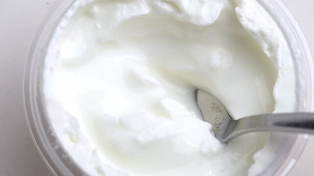 Spoon scooping thick white Greek yogurt from a plastic container. Top view of creamy dairy product texture. Concept of healthy breakfast