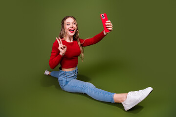 Fototapeta premium Happy young woman taking a selfie while sitting on a green studio background wearing a red top and jeans