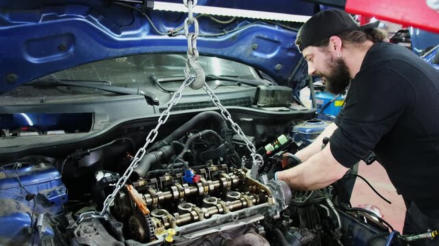 Mechanic disconnects car engine hanging on hoist in repair shop slow motion. Bearded technician prepares vehicle motor for maintenance at station