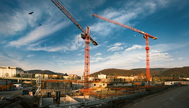 Large construction site with red cranes looming into the blue sky with dramatic clouds. Wide angle tilted up view. 