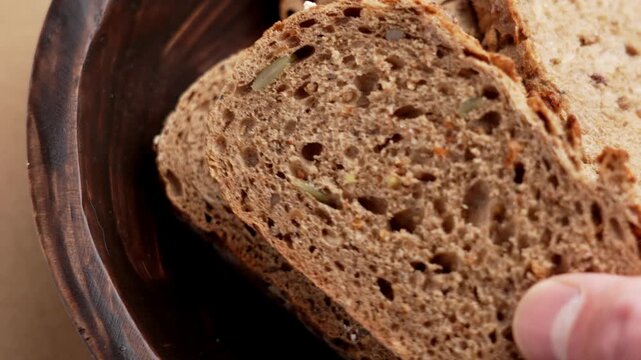 Close up of human hand taking a slice of dark German bread from a wooden bowl with whole grain crumb and seeds inside. Rustic baked product