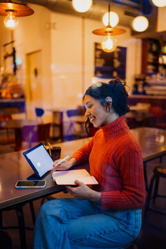 Side view of smiling hispanic woman working remotely with tablet and paper diary in coffee shop at night, distance learning student taking notes, freelance lifestyle, professional occupation.