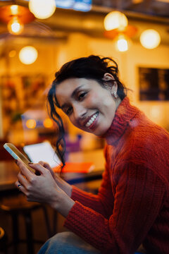 Cheerful mixed race female looking at camera while holding smartphone in coffee shop, social media influencer, mobile banking and chatting online, authentic lifestyle, looking at camera.
