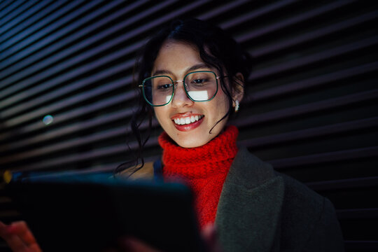 Happy multiracial woman in glasses browsing tablet content outdoors, digital communication and online shopping, tech savvy professional on nighttime city street, authentic representation.