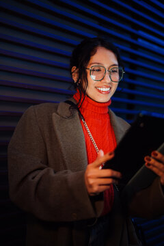 Happy mixed race female using digital tablet outdoors in evening city lights, scrolling mobile app and smiling, creative student learning online, tech savvy lifestyle, authentic representation.