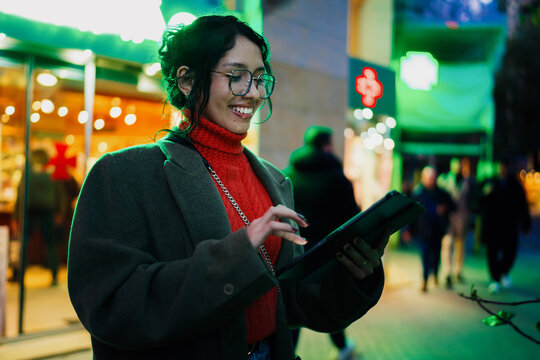 Smiling mixed race woman using digital tablet on busy city street with neon lights, mobile application user, remote work and freelance lifestyle, tech savvy professional, night city.