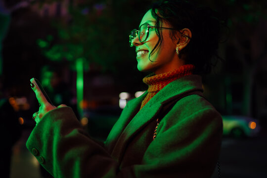 Side view of smiling mixed race woman using smartphone on night city street with green neon lights, authentic representation of digital nomad, mobile networking and communication concept.