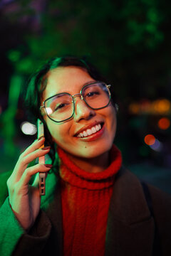 Confident mixed race woman in glasses holding smartphone and smiling, authentic representation of ethnic millennial, positive attitude and confidence, urban nighttime with green light.
