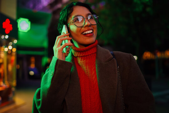 Cheerful multiracial woman in glasses talking on smartphone outdoors at night under green lighting, authentic representation of young professional communicating, mobile office concept.