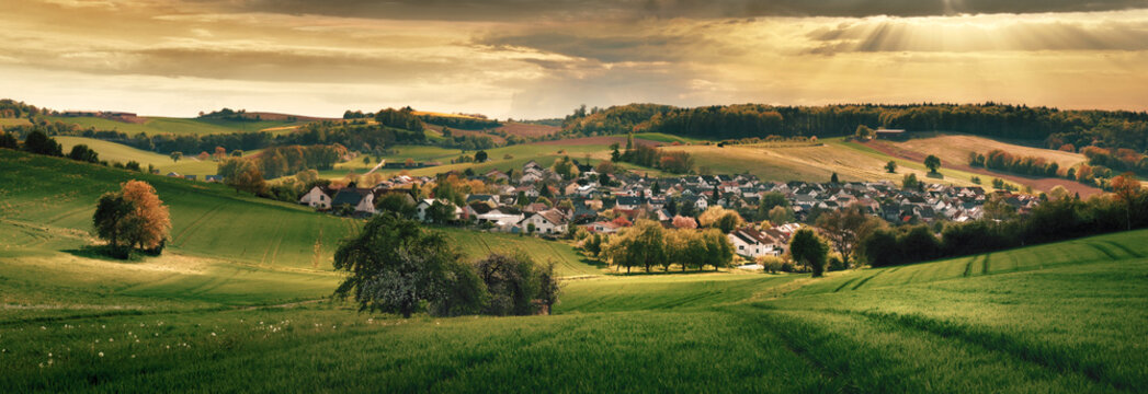 Village in a valley in golden dramatic light. A panoramic tranquil rural scenery with a scenic small town surrounded with green meadows on hills 
