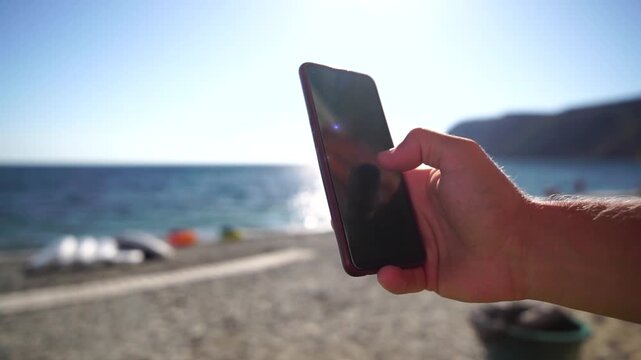 Man sea smartphone. The young man in sunglasses, sunbathing by the sea and spending time on his mobile phone texting with girlfriend, looks at the camera in surprise, sees a news on the phone.
