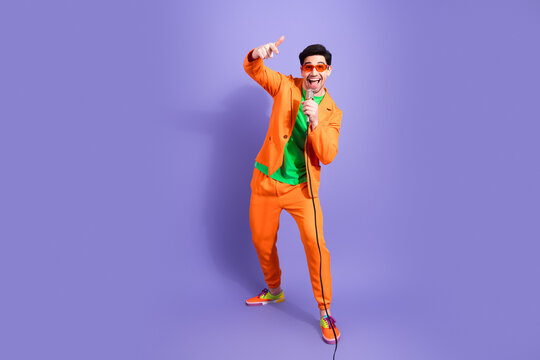 Cheerful singer in orange suit performing with microphone and giving thumbs up against purple studio background, colorful pop entertainer smiling and energetic during a fun music performance