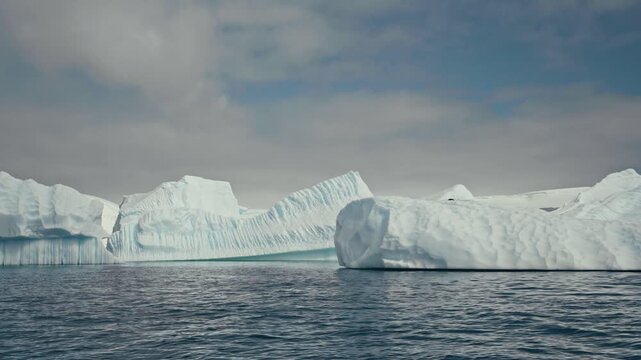 Slow pan along a row of massive striated tabular icebergs with eroded vertical walls and rounded bulging formations above dark rippling Antarctic polar ocean waters