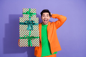 Obraz premium Surprised young man holding a tall stack of wrapped Christmas gifts in bright orange jacket and green shirt against a purple studio background