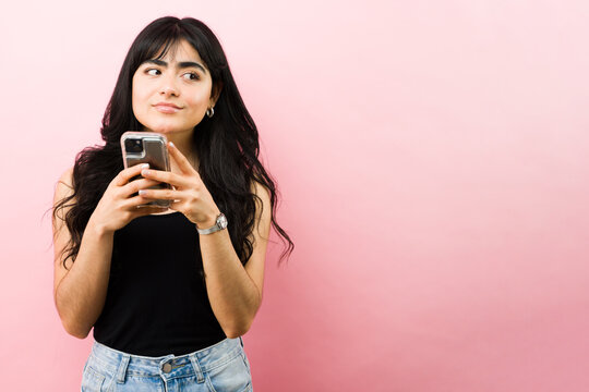 Young latin woman thinking, holding smartphone, looking away in doubt, standing on pink background