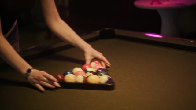 Tilt down shot of young adult woman setting up billiard table with pool balls then removing rack before start of game at low key lit bar