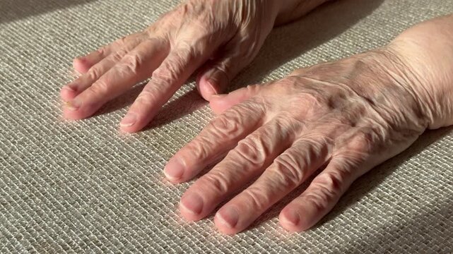 Elderly hands close-up on fabric, sunlight highlighting wrinkles and veins, fingers gently splayed, nails trimmed, soft shadow