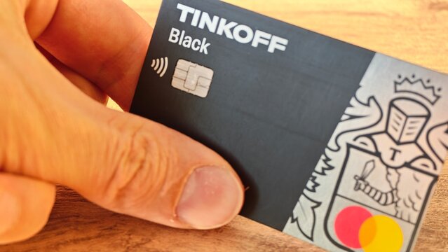 Sosnoviy Bor, Russia - April 21, 2026: Hand holding Tinkoff Black contactless payment card with Mastercard logo on wooden surface, showcasing modern banking technology