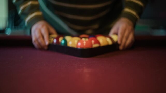 Rack focus close up shot of unrecognizable man racking set of pool balls into triangle on red billiard table while placing five number ball at front