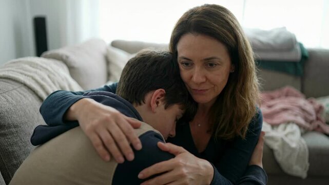 Mother comforting teenager boy in living room. Parent hugging struggling child on couch. Connectioneering concept of emotional support during difficult moment home.