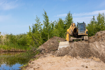 A yellow tracked excavator works along a wetland pond bank, moving soil and clearing vegetation for land development and erosion control. Bright blue sky and green trees frame the industrial scene © evannovostro