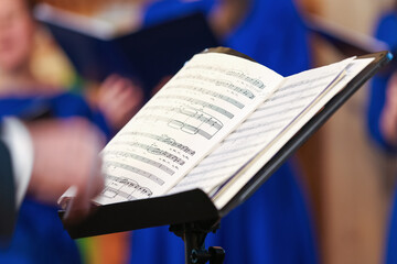 Naklejka premium Open sheet music on a stand as a conductor leads a choir in blue robes, capturing focused singers, harmony and the intimate atmosphere of a choral rehearsal or live vocal performance