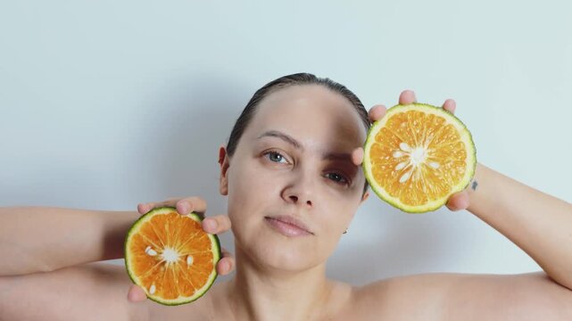 Woman holding two vibrant citrus halves at face level against a light background, suggesting vitamin c skincare, natural beauty, glowing healthy complexion and refreshing self care routine