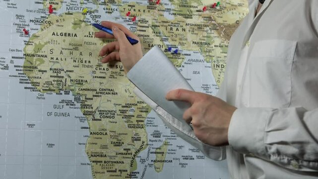 Close-up of a man holding a notebook and pen, standing in front of a world map, studying geography, where to go and writing down his thoughts in a notebook