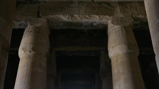 Mysterious dark corridor between colossal carved columns inside ancient Abydos temple, Egypt, with hieroglyphic reliefs, painted ceiling fragments and dramatic spot lighting.